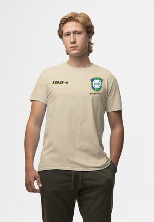 Jeune homme aux cheveux blonds portant un t-shirt beige avec « 1994 » et l'emblème de football du Brésil CBD, debout devant un fond blanc uni.