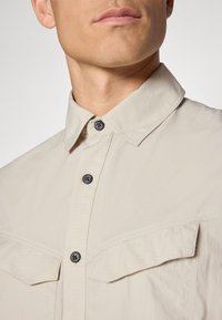 Lichtbeige button-up shirt met twee borstzakken, een kraag en zwarte knopen. De stof heeft een subtiele textuur.