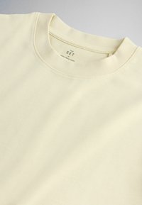 T-shirt in cotone beige chiaro con scollo rotondo, cuciture visibili sul colletto e etichetta stampata con taglia e origine all'interno del collo.