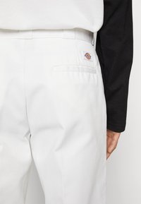 Pantaloni di cotone bianchi con una sola tasca posteriore, caratterizzati da un piccolo logo Dickies multicolore. Indossati con una camicia nera a maniche lunghe.