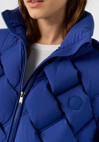 Blaue Puffjacke mit gestepptem Muster, hohem Kragen und einem Reißverschluss vorne. Verfügt über ein Logo-Abzeichen auf der linken Brustseite.