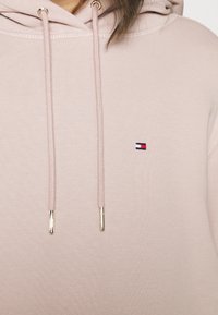 Sudadera rosa claro hecha de una tela suave con una superficie texturizada, que presenta un pequeño logo bordado y extremos de cordón con acentos dorados.