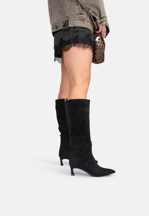 Stivali alti neri in suede con punta affusolata e tacco sottile. Indossati con pantaloni corti neri con pizzo e una giacca di jeans.