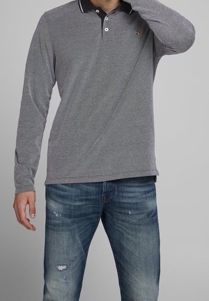 Mann trägt ein graues Polohemd mit langen Ärmeln und ausgewaschene blaue Jeans mit abgenutzten Details, steht vor einem einfarbigen Hintergrund.