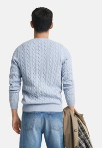 Helles blaugraues Strick-Sweatshirt mit gerippten Bündchen und Saum, kombiniert mit blauen Jeans. Das Model hält eine beige Jacke.