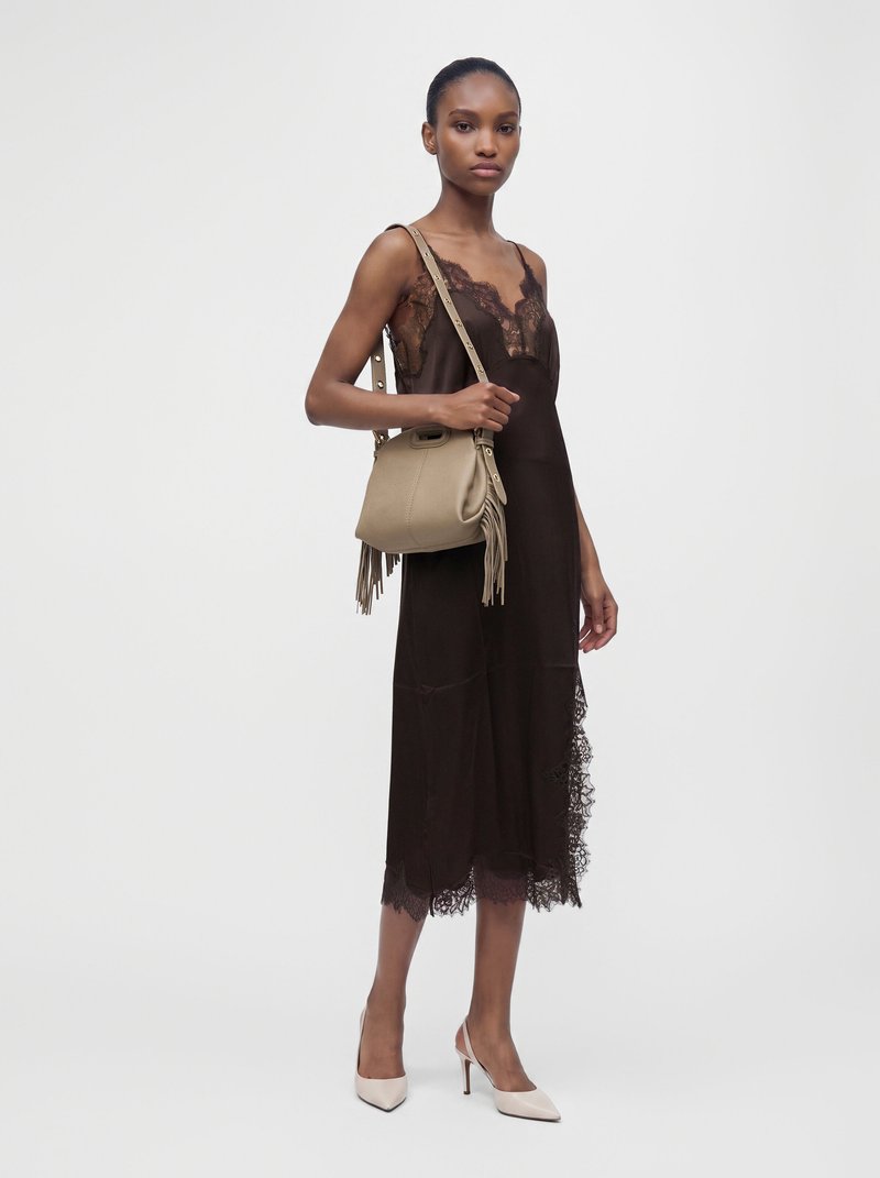 Femme portant une robe noire en dentelle sans manches, un sac bandoulière beige avec des franges, et des chaussures blanches à talons hauts, debout sur un fond uni.