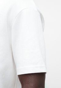 Κοντινό πλάνο σκούρου δέρματος χεριού που φοράει λευκό κοντομάνικο πουκάμισο σε απλό φωτεινό φόντο.