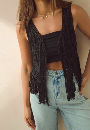 Veste noire perlée avec un ourlet à franges, présentant un design en maille ouverte texturée. Associée à un crop top noir et un jean taille haute bleu clair.
