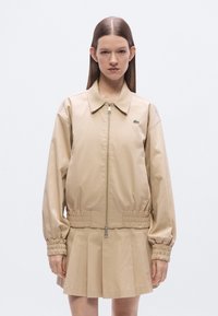 Μπεζ μπουφάν τύπου bomber με φερμουάρ μπροστά, ελαστική μέση και μανίκια με σούρες. Διαθέτει μικρό πράσινο λογότυπο στο αριστερό στήθος.