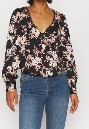 Kvinde iført en sort blomsterbluse med knapper foran og lange, puff-ærmer, kombineret med højhængte blå jeans med knappelukning.