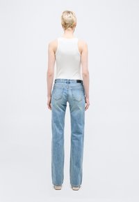 Άτομο με κοντά ξανθά μαλλιά, φορώντας λευκό αμάνικο τοπ και ανοιχτό μπλε τζιν με ευθείες γραμμές, στέκεται γυρισμένο προς τα πίσω σε απλό φόντο.