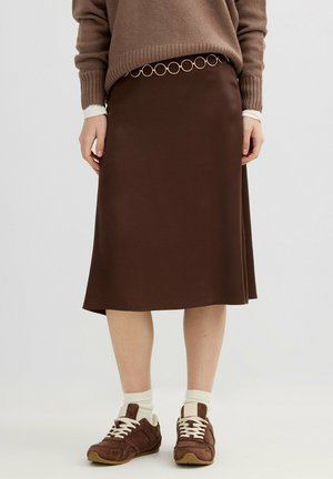 Mujer con falda midi marrón, suéter beige y zapatillas marrones con cordones blancos y calcetines, de pie frente a un fondo blanco liso.