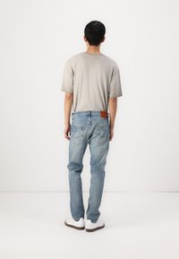 T-shirt en coton gris, jean en denim bleu clair avec une patch en cuir marron, baskets blanches, vues de derrière sur un fond uni.