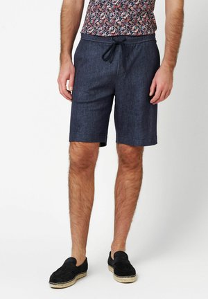 Marineblaue Shorts mit Kordelzugbund, gefertigt aus einem denimähnlichen Stoff. Verfügt über eine legere Passform und gesäumte Kanten. Schwarze Slipper.