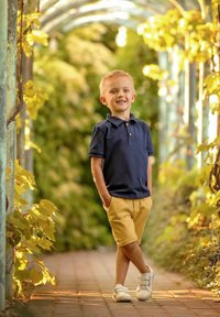 Dziecko w granatowej koszulce polo i żółtych szortach stoi na chodniku, otoczone zielenią, z rękami w kieszeniach i uśmiechem. Białe trampki.