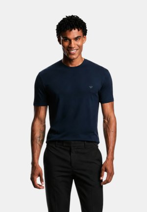 Uomo sorridente con capelli corti e ricci che indossa una t-shirt aderente blu navy e pantaloni neri, in piedi davanti a uno sfondo chiaro uniforme.