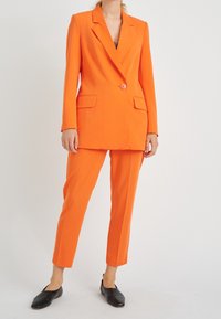 Oranje op maat gemaakte blazer en bijpassende broek van soepele stof. De blazer heeft een enkele knop en twee voorkruiszakken. Zwarte instappers.