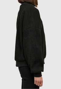 Pullover in felpa nera con superficie testurizzata, vestibilità oversized e polsini a costine; presenta un colletto alto e spalle scese.