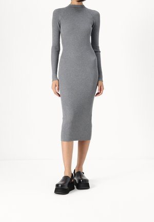 Femme portant une robe grise côtelée ajustée, à manches longues, longueur genou, avec des chaussures à plateformes noires, debout sur fond blanc.
