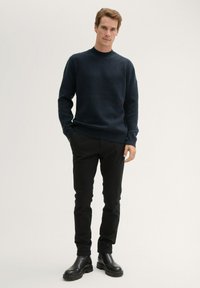 Pull en maille bleu marine avec col rond, associé à un pantalon noir slim et des bottines en cuir noir à la cheville. Texture lisse, design ajusté.