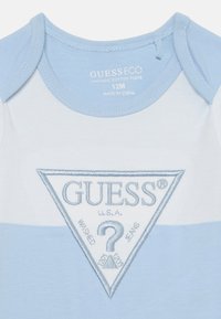 Un body pour bébé en coton organique bleu clair avec un logo brodé en argent en forme de triangle, présentant le nom de la marque "GUESS" et un détail de point d'interrogation.
