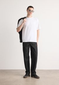 Hvit t-skjorte med korte ermer kombinert med mørk denim bukser og grå sko. Modellen holder en svart jakke over den ene skulderen og har solbriller på.