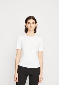 Tricou alb cu mânecă scurtă, gât circular, textură netedă, design ajustat și cusături minime, purtat cu pantaloni negri. Fără modele vizibile.