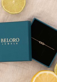 Goldenes Unendlichkeit-Armband in einem schwarzen Samtinneren einer türkisfarbenen Box mit der goldenen Aufschrift "BELORO JEWELS", umgeben von Zitrusscheiben.