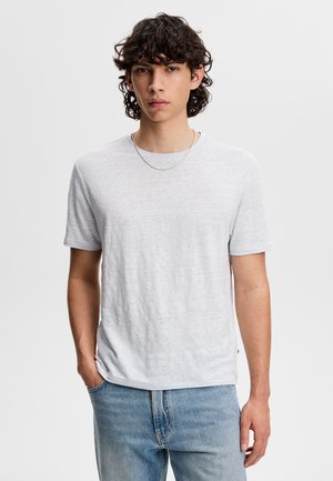Junger Mann mit lockigem dunklem Haar, der ein schlichtes weißes kurzärmeliges T-Shirt, hellblaue Jeans und eine dünne silberne Halskette trägt.