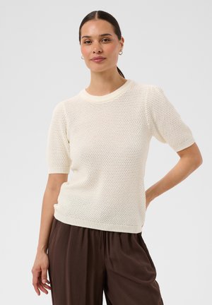Femme portant un pull texturé crème à manches courtes et un pantalon marron ample, debout avec une main sur la hanche devant un fond uni.