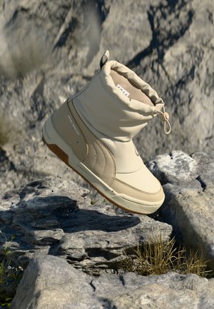 Botte imperméable beige avec un col rembourré, une semelle en caoutchouc et un design sans coutures, reposant sur un terrain rocheux avec de l'herbe en arrière-plan.