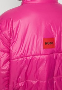 Chaqueta acolchada en rosa brillante con una textura lisa y brillante. Presenta una etiqueta rectangular roja con la palabra "HUGO" en negro. Se pueden ver costuras detalladas.