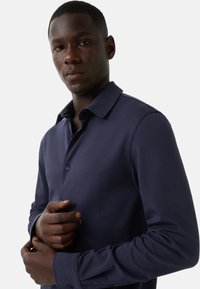 Man in een donkerblauw overhemd met lange mouwen, die een manchet aanpast met een neutrale expressie tegen een effen achtergrond.