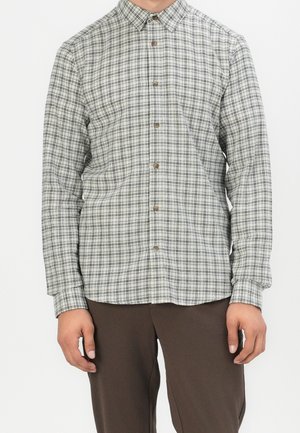 Mężczyzna ubrany w białą koszulę na guziki z czarnym wzorem w krate oraz brązowe spodnie, stojący na tle jednolitej białej ściany.