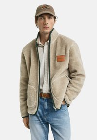Beige fleecejack met een groene rits en accent, voorzien van twee grote zakken en een leren embleem met het opschrift "GANT." Draagt over een geruit shirt.