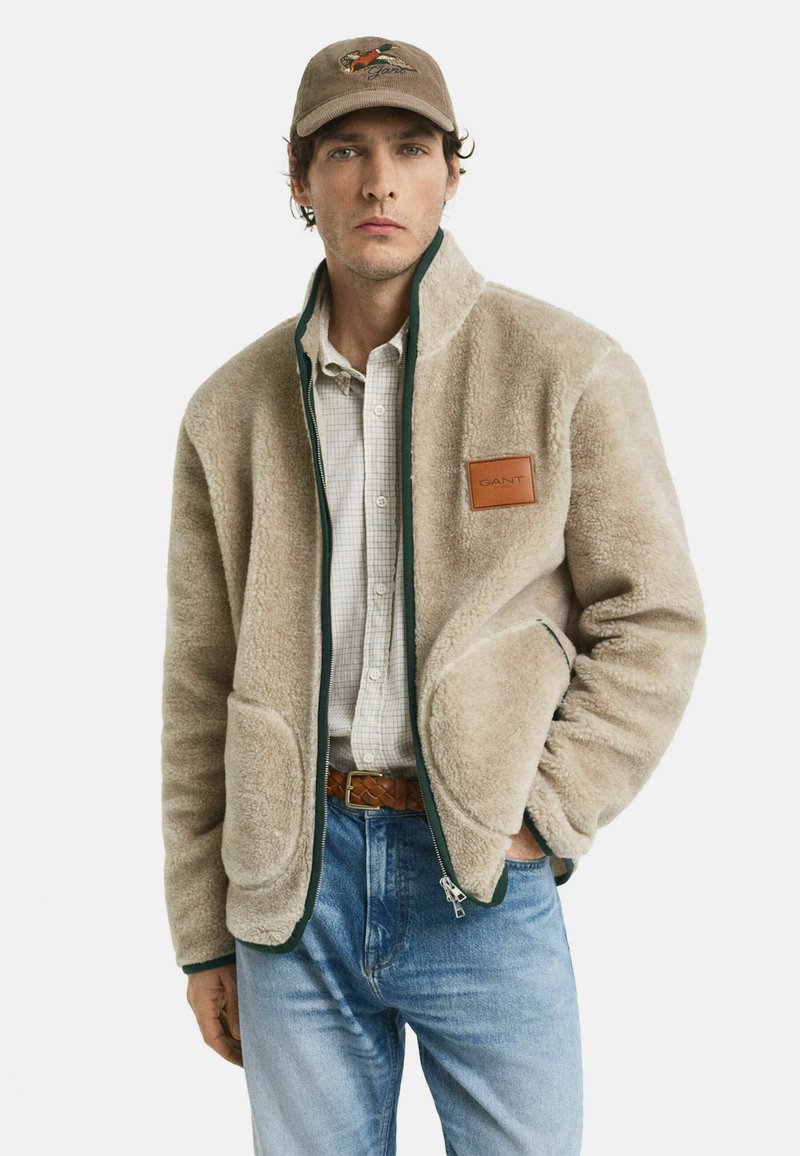 Beige fleecejack met een groene rits en accent, voorzien van twee grote zakken en een leren embleem met het opschrift "GANT." Draagt over een geruit shirt.
