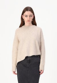 Beige pulover iz volnene mešanice s širokim izrezom, spuštenimi rameni in skrajšanim robom, v kombinaciji s črnimi jeans kratkimi hlačami.