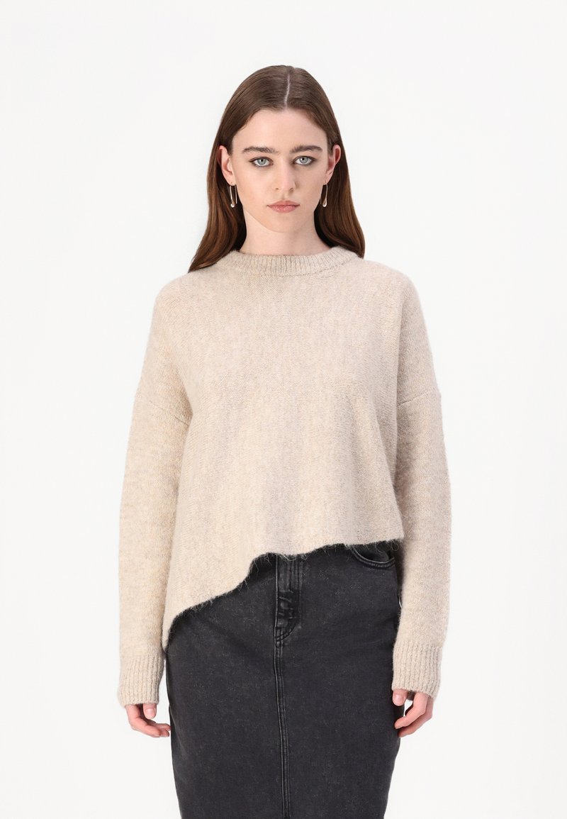 Beige pulover iz volnene mešanice s širokim izrezom, spuštenimi rameni in skrajšanim robom, v kombinaciji s črnimi jeans kratkimi hlačami.