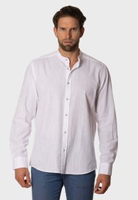 Camisa blanca de manga larga con cuello alto, cierre de botones y una textura sutil; presenta un pequeño logo en el pecho. Llevada con pantalones azules.