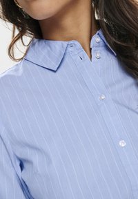 Woman wearing a light blue pinstriped button-up shirt with a classic collar and white buttons.