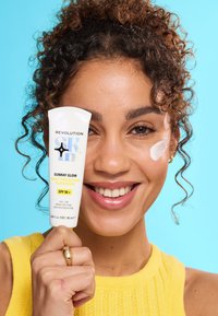 Un tube de crème solaire étiqueté "Sunray Glow Protection Quotidienne SPF 50+" en blanc avec des accents colorés, tenu par une personne, avec une touche sur sa joue.