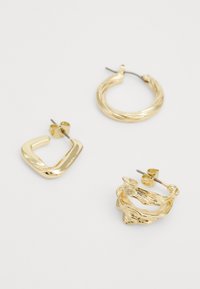 Tři páry zlatých náušnic: čtvercové, zkroucené kruhy a textura kruhů s organickými tvary a lesklými povrchy.