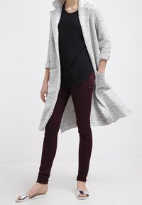 Harmaa teksturoitu pitkä takki, musta toppi, viininpunaiset kapeat housut ja hopeiset liu'utettavat sandaalit. Takissa on kaulus ja avoin etuosa.