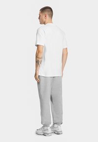 Biała koszulka z krótkim rękawem, szare dresy i białe sneakersy. Gładki materiał, luźny krój, z widocznymi tatuażami na ramieniu.