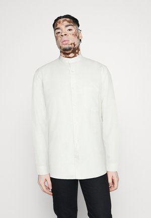 Camisa de manga larga color blanco roto con cuello mao, cierre de botones y un bolsillo en el pecho. Textura suave, corte relajado.