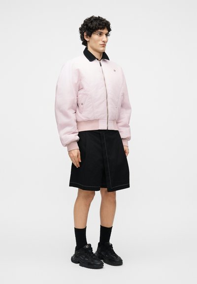 Νέος με σγουρά μαλλιά, φορώντας ροζ ανοιχτό μπουφάν τύπου bomber, μαύρη φούστα μέχρι το γόνατο, μαύρες κάλτσες και μαύρα αθλητικά, στέκεται πλάγια.