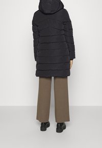 Abrigo acolchado azul marino con capucha y diseño acolchado horizontal. Combinado con pantalones anchos marrones y botas negras gruesas.