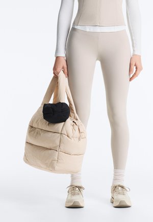 Henkilö, jolla on beigenväriset leggingsit ja lenkkarit, pitää kädessään suurta beigenväristä toppattua kangaskassia, johon on kiinnitetty pieni musta pussi.