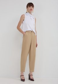 Blusa bianca senza maniche con dettaglio a fiocco, abbinata a pantaloni beige a vita alta. La modella indossa scarpe col tacco color bordeaux. Sfondo neutro.