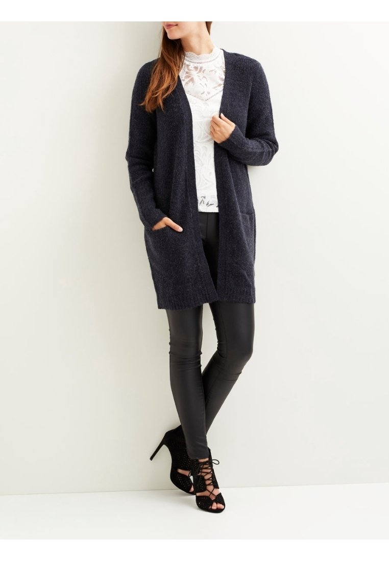 Femme portant un long cardigan sombre, un haut en dentelle blanche, un pantalon en cuir noir et des chaussures à lacets à talons hauts noires, debout contre un mur blanc.